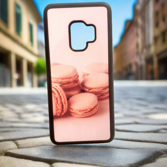 A smartphone case with a picture of pink macarons sits upright on a out door  backdrop.